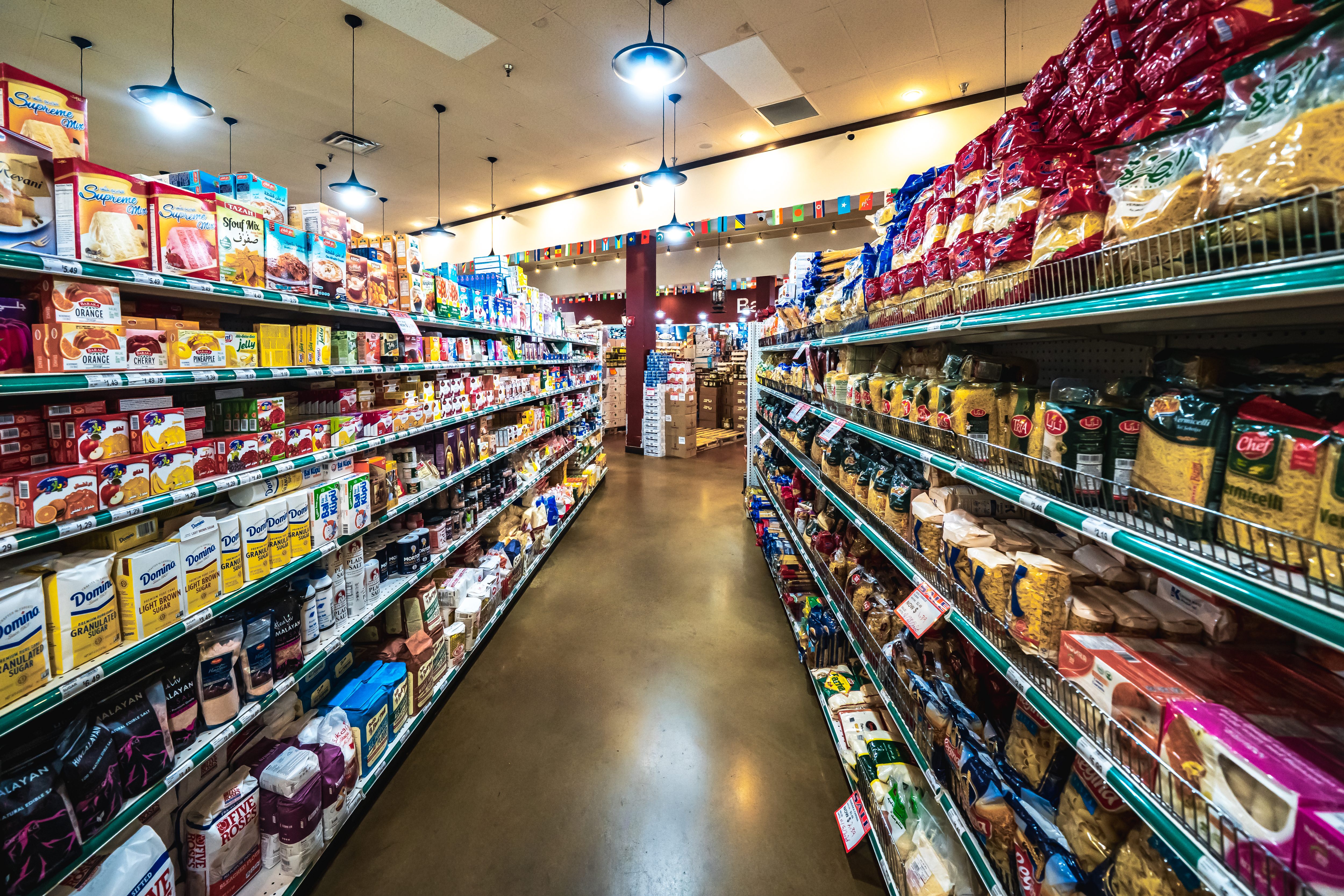 A wide aisle in a grocery store stocked with packaged food items like pasta, sugar, and baking mixes on both sides.