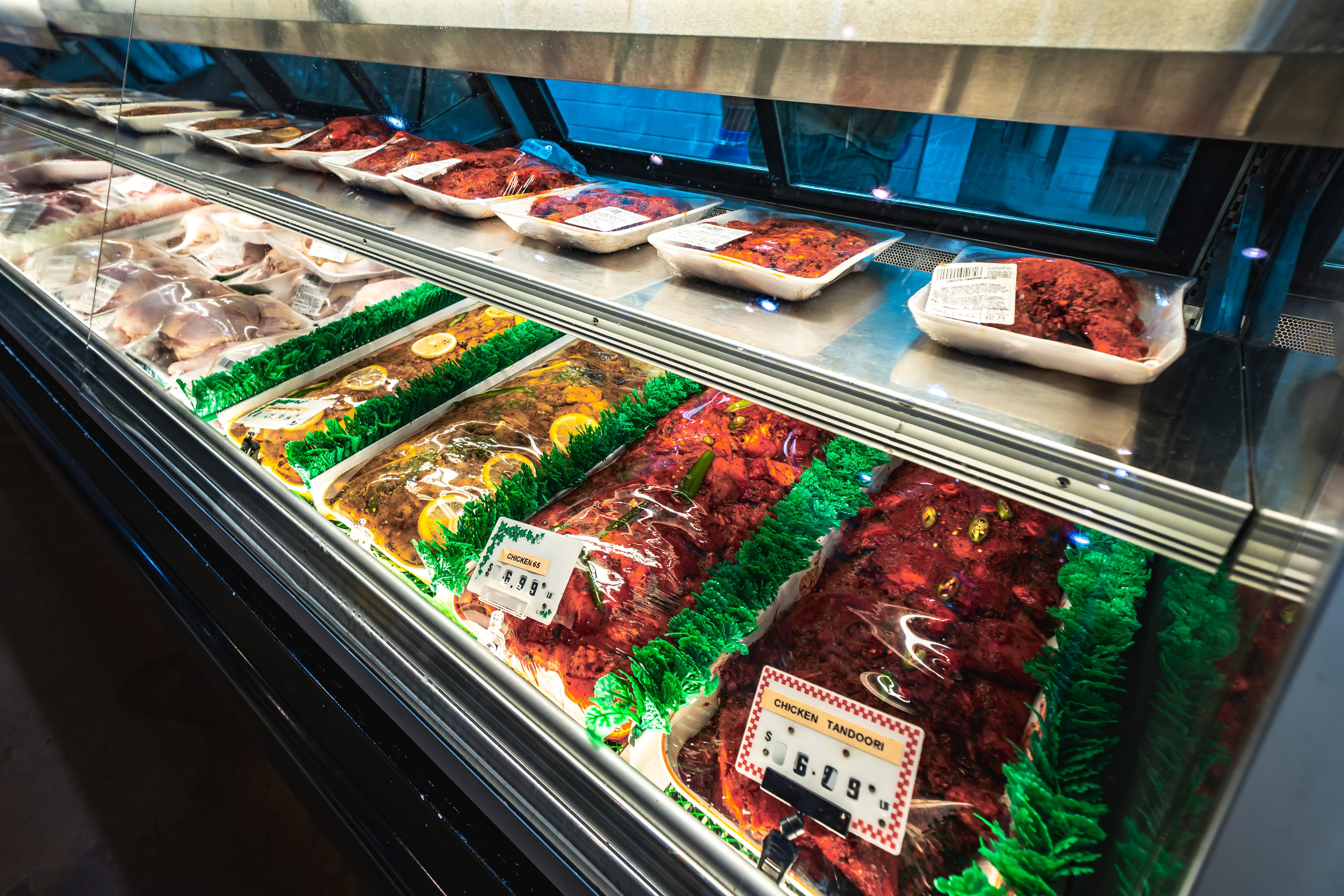 Glass display case in a market showing trays of marinated meats including chicken tandoori and chicken 65, with price tags.