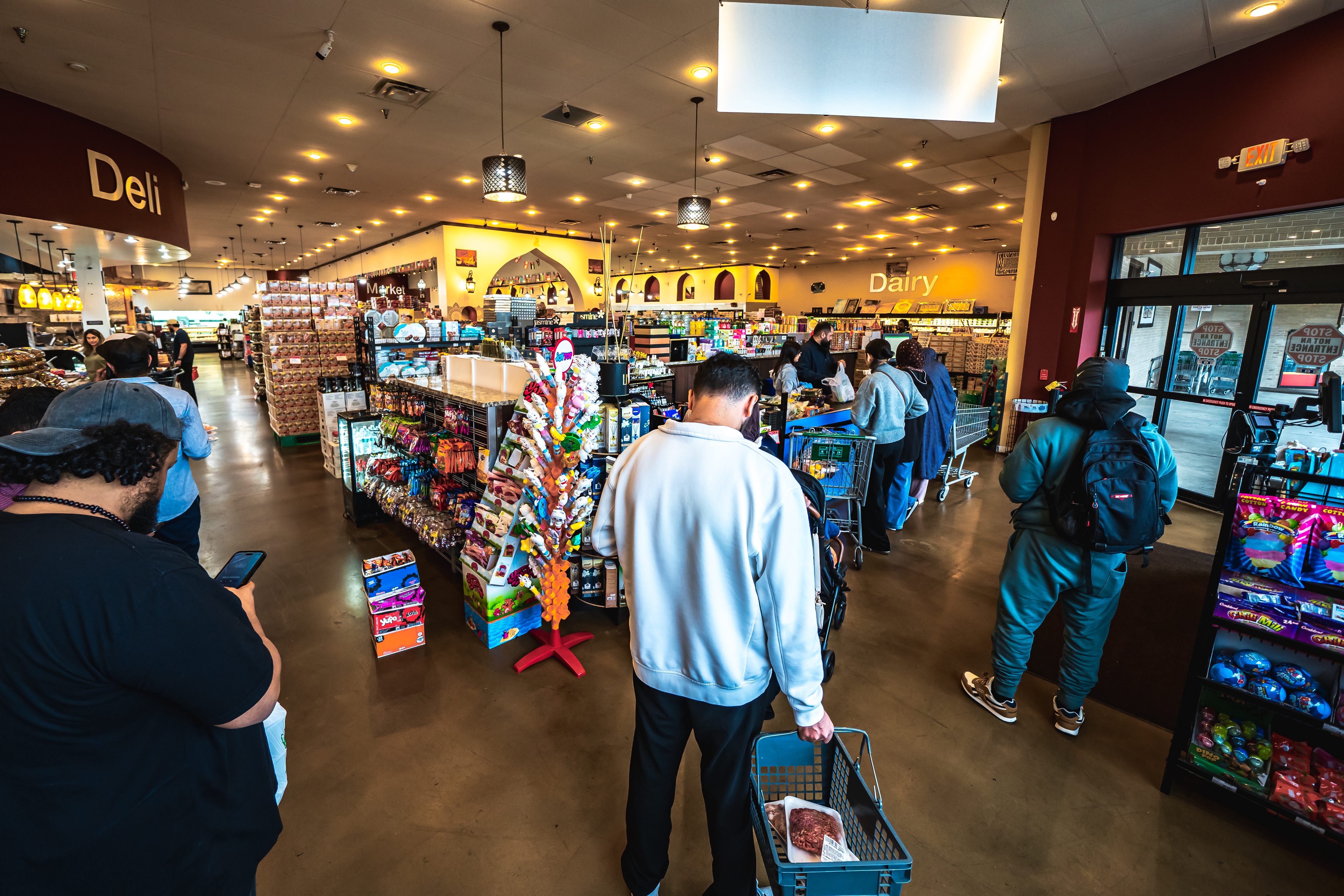 People waiting in line with shopping baskets and carts inside a well-lit grocery store near deli and dairy sections.