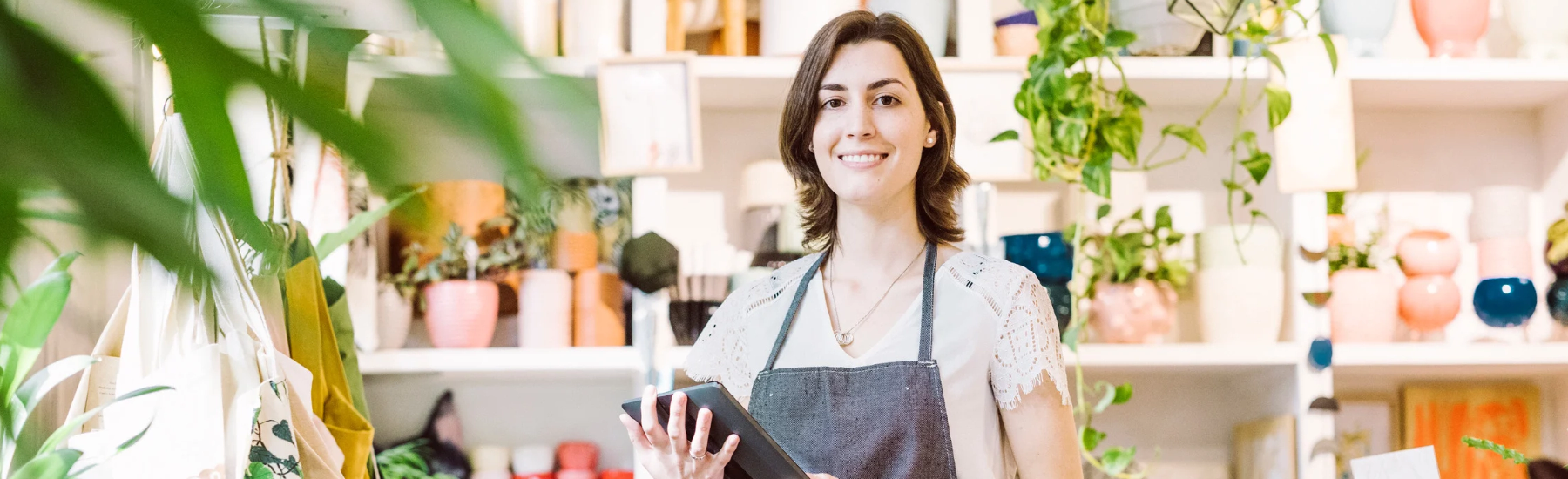 Smiling woman in an apron holding a tablet inside a bright plant and pottery store.