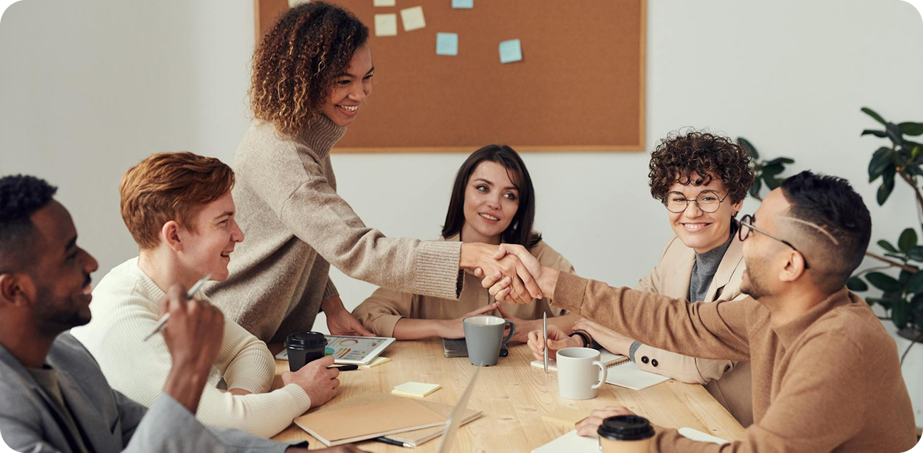 Group of diverse colleagues sitting around a table, two women smiling and shaking hands during a meeting.