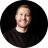 Smiling man with light skin and short hair wearing a black hoodie against a black background.
