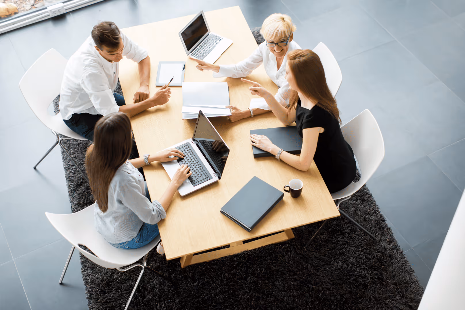 Four people sitting around a wooden table in an office, working with laptops, tablets, documents, and discussing ideas.