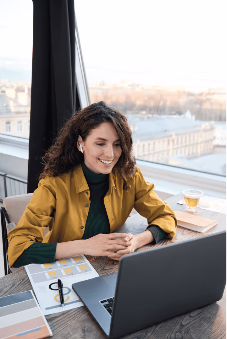 Smiling woman with curly hair wearing yellow jacket and green sweater, sitting at a desk with laptop, papers, and tea cup near window.
