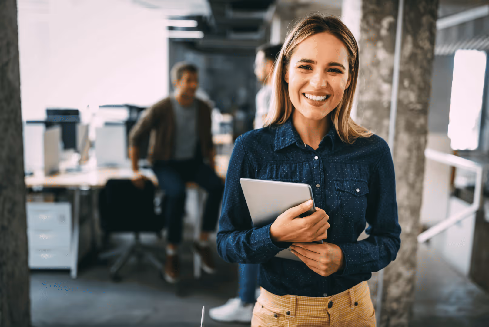 Smiling woman holding a tablet standing in a modern office with two blurred colleagues in the background.