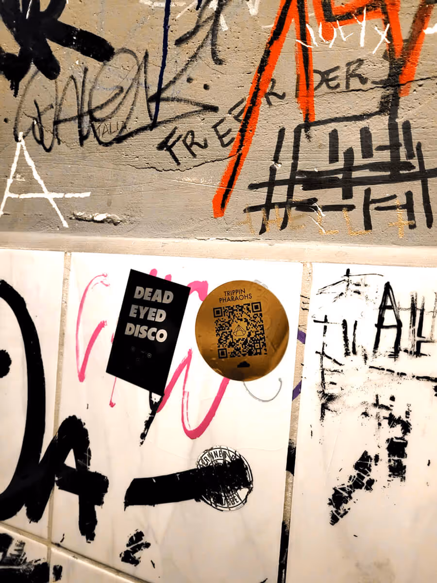 Wall with graffiti and two stickers, one reading 'DEAD EYED DISCO' and a round gold 'TRIPPIN PHARAOHS' QR code sticker.