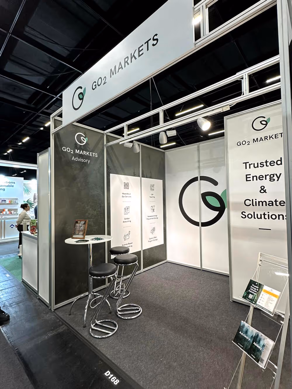 Exhibition booth for GO2 MARKETS with white and gray panels, bar stools, and posters about energy and climate solutions.
