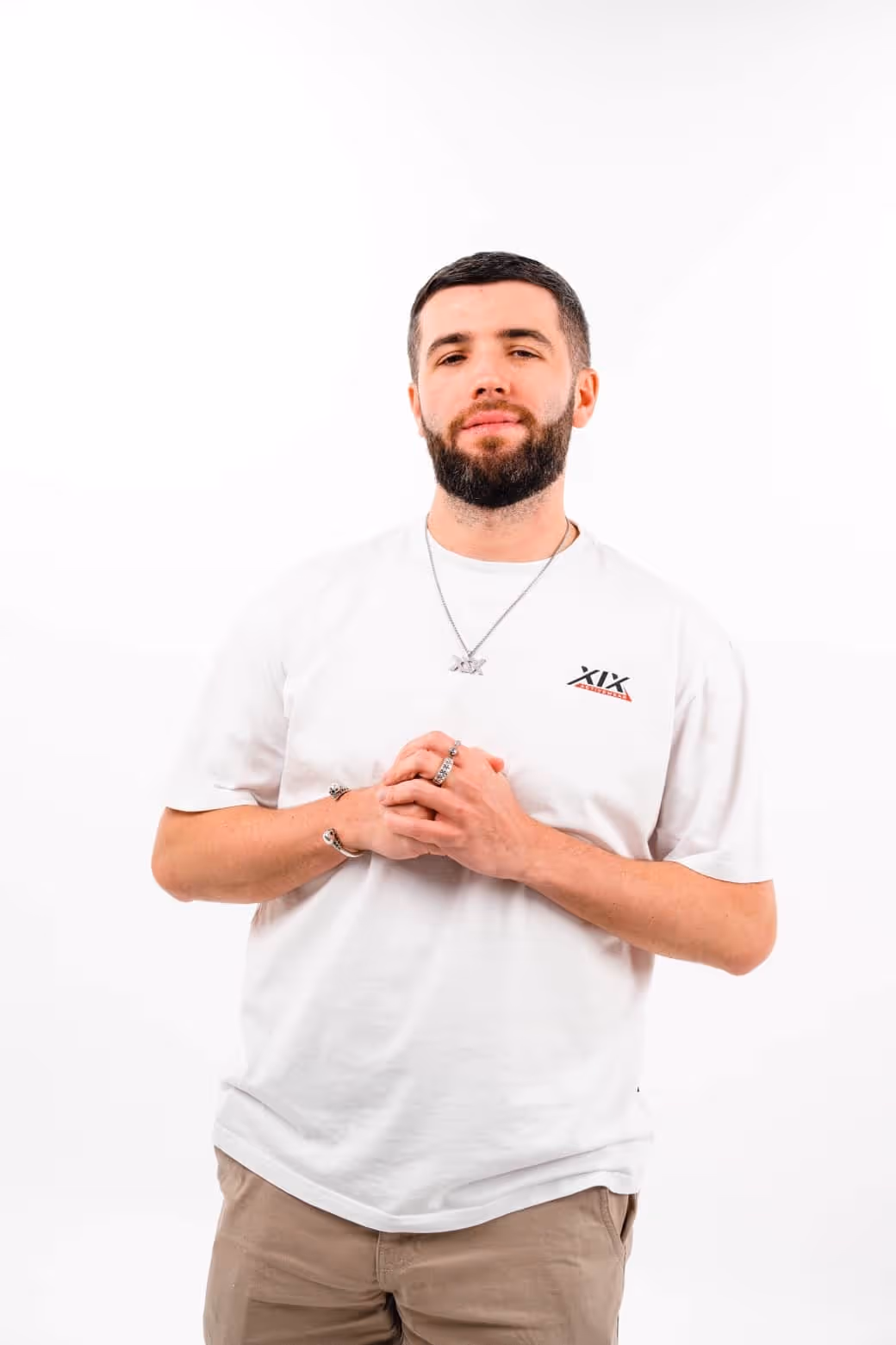Bearded man clasping hands wearing a white XIX Athletics t-shirt and beige pants standing against a white background.