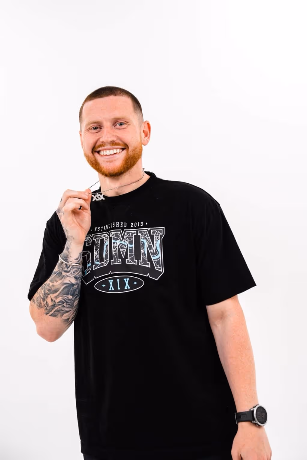 Smiling man with a beard and tattooed arm wearing a black graphic t-shirt holding a necklace pendant near his face.