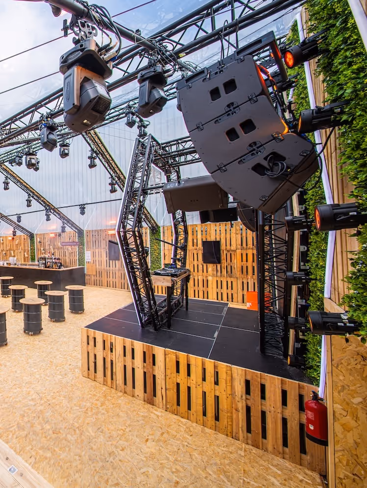 Scène de DJ vide avec éclairage et haut-parleurs suspendus dans une salle à toit transparent avec murs en palettes de bois et tabourets cylindriques.