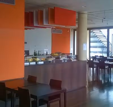 Salle de cantine moderne avec un comptoir de buffet, des tables et des chaises en bois sombre, et un mur orange vif.