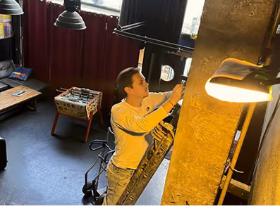 Un homme sur une échelle ajuste un projecteur dans une pièce avec rideaux rouges et baby-foot.