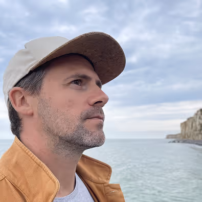 Homme portant une casquette beige et une veste marron regardant vers la mer avec un ciel nuageux en arrière-plan.