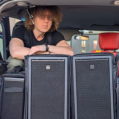 Jeune homme aux cheveux bouclés et lunettes s'appuyant sur de grosses enceintes dans le coffre d'une voiture.