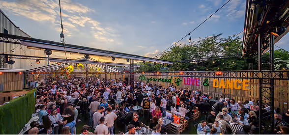 Grande foule rassemblée dans un espace extérieur décoré avec une enseigne indiquant « All you need is LOVE & DANCE » sous un ciel au crépuscule.
