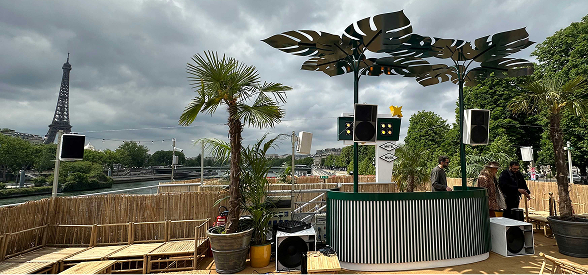 Espace extérieur avec un bar contemporain sous un ciel nuageux, palmiers, la tour Eiffel et la Seine en arrière-plan.