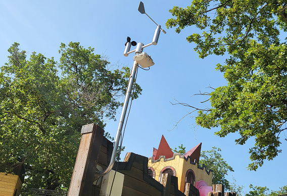 Capteur de surveillance météorologique et sonore installé sur un poteau en bois dans un parc avec des arbres verts et un château de jeu coloré en arrière-plan.