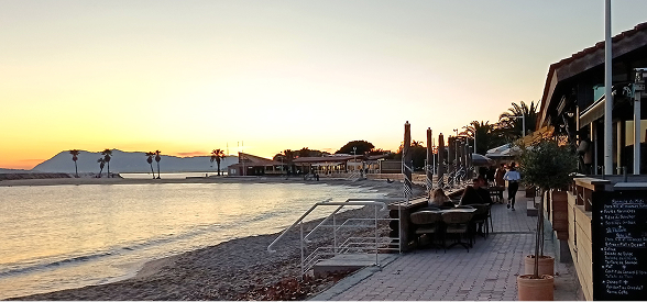 Plage au coucher du soleil avec palmiers, tables de restaurant en terrasse sur une promenade pavée.
