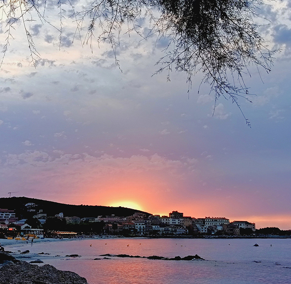 Coucher de soleil coloré sur une ville côtière avec des collines, la mer calme au premier plan et des branches d'arbre au sommet.