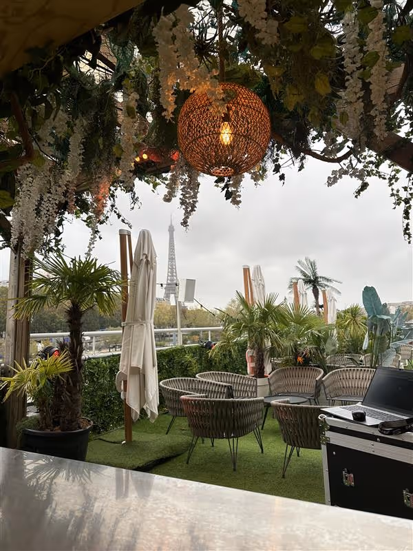 Terrasse extérieure avec chaises en osier, plantes en pot et lampes suspendues, avec vue sur la tour Eiffel par temps nuageux.