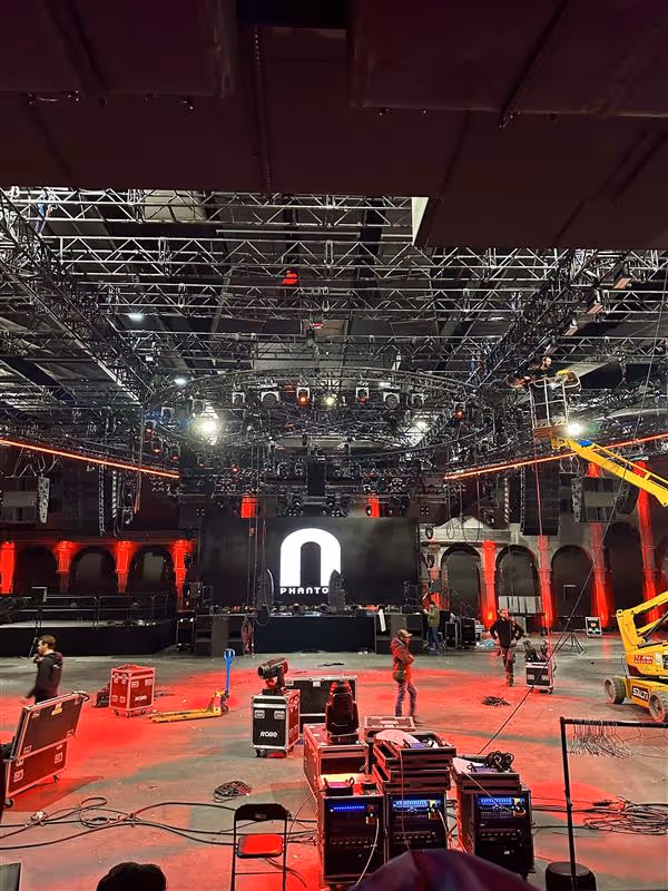Installation en cours de la scène de concert sur le thème du rouge et du noir. Les techniciens ajustent le matériel sous les lumières vives de la scène et les structures métalliques.