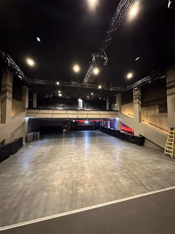 Une grande salle de concert vide avec un sol gris, des gradins, des projecteurs suspendus au plafond et une échelle jaune appuyée contre le mur de droite.