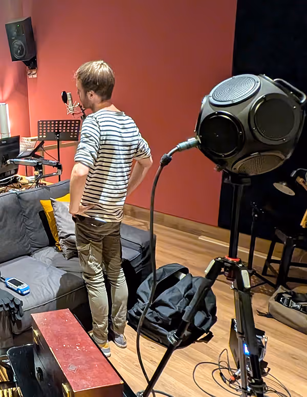 Personne debout devant un micro dans un studio d'enregistrement avec un ingé son assis derrière un bureau équipé d'ordinateurs.