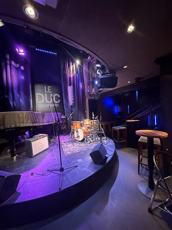 Scène de jazz intime avec batterie, piano couvert, amplificateur et microphones dans un club nommé Le Duc des Lombards.