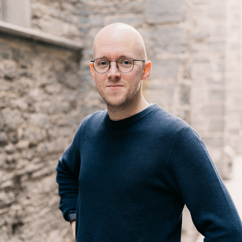 Man with glasses wearing a dark blue sweater standing outdoors against a stone wall.