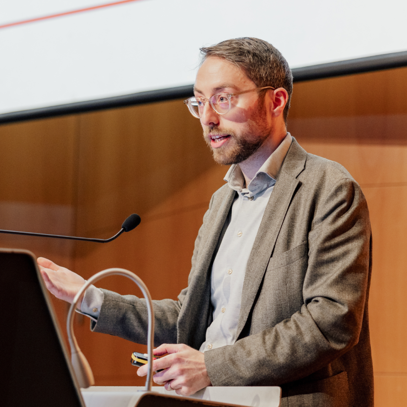 Man with glasses and beard speaking at a podium with a microphone in a lecture hall.