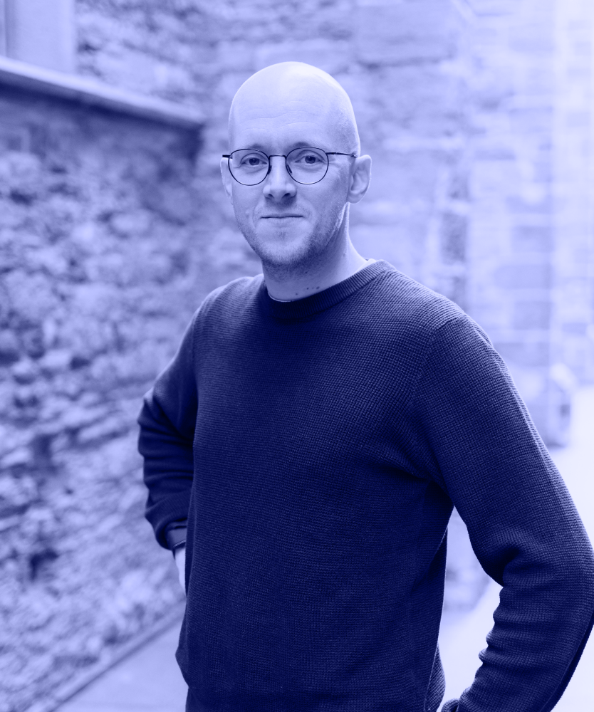 man with glasses wearing a navy blue sweater standing outdoors near a stone wall.
