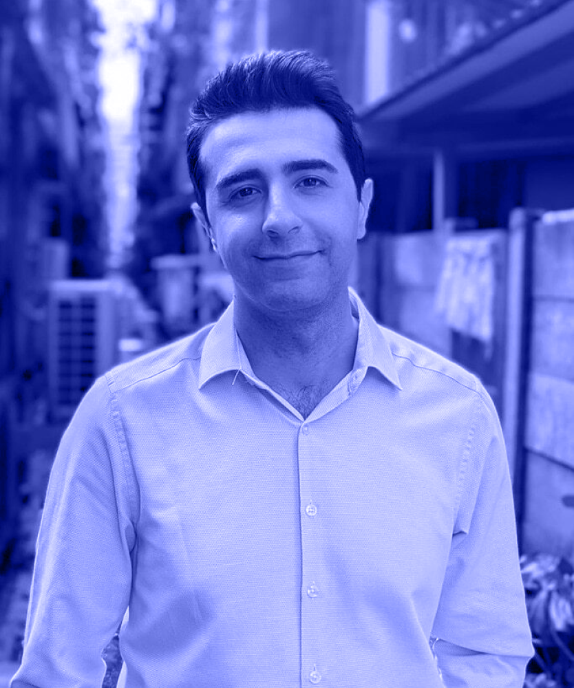 Smiling man with dark hair wearing a light blue button-up shirt standing outdoors in an alley.