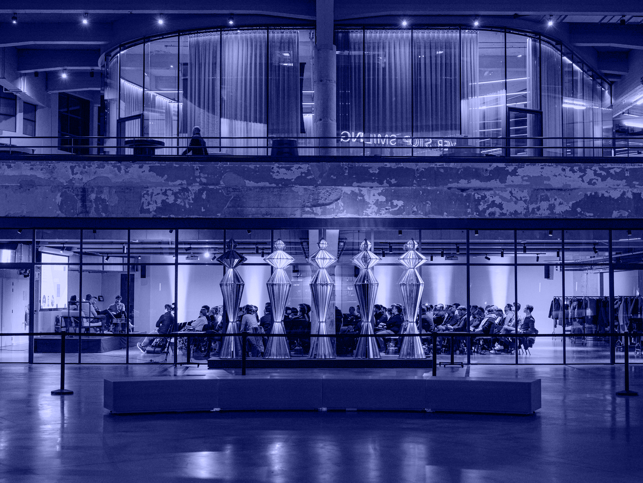 Indoor modern space with seated audience watching a panel discussion behind glass walls and tall metallic sculptures in the foreground.