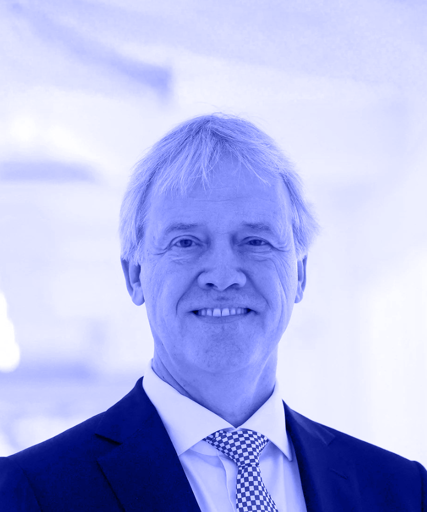 man with glasses wearing a navy blue sweater standing outdoors near a stone wall.