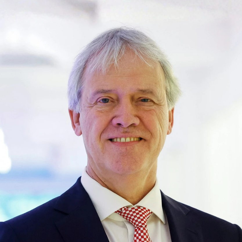 Man with glasses wearing a dark blue sweater standing outdoors against a stone wall.