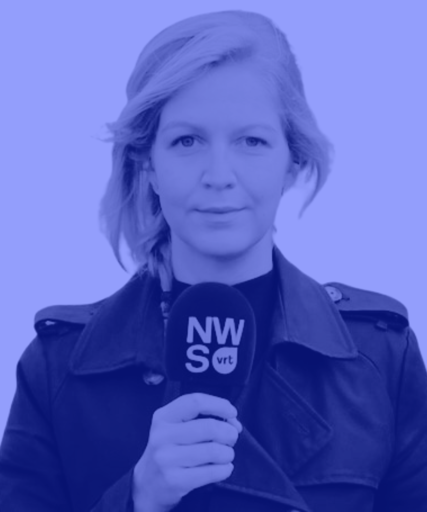 man with glasses wearing a navy blue sweater standing outdoors near a stone wall.