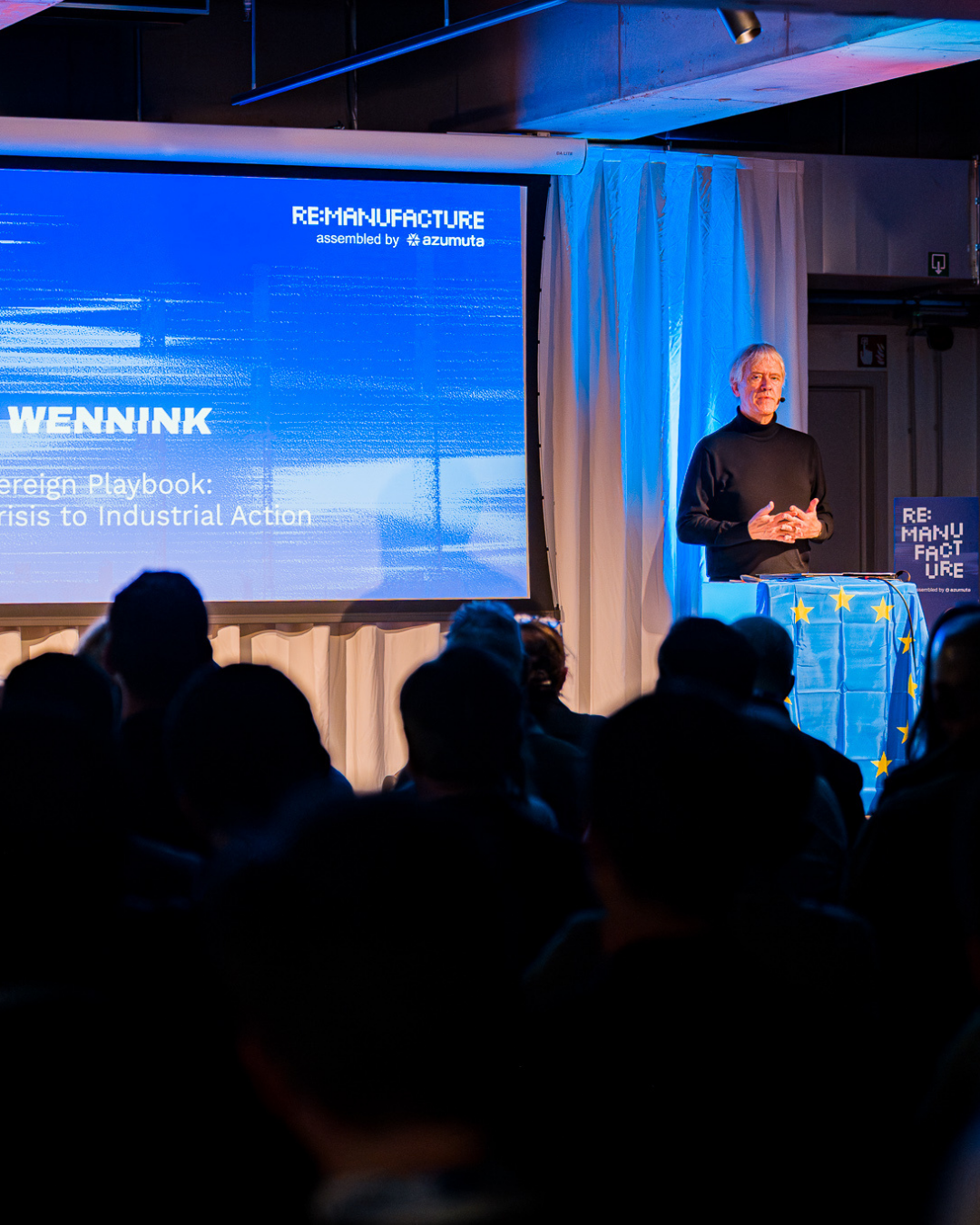Indoor modern space with seated audience watching a panel discussion behind glass walls and tall metallic sculptures in the foreground.