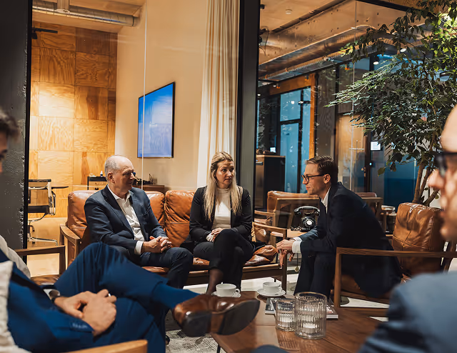 A group of five people in business attire sitting in a modern office lounge having a discussion.
