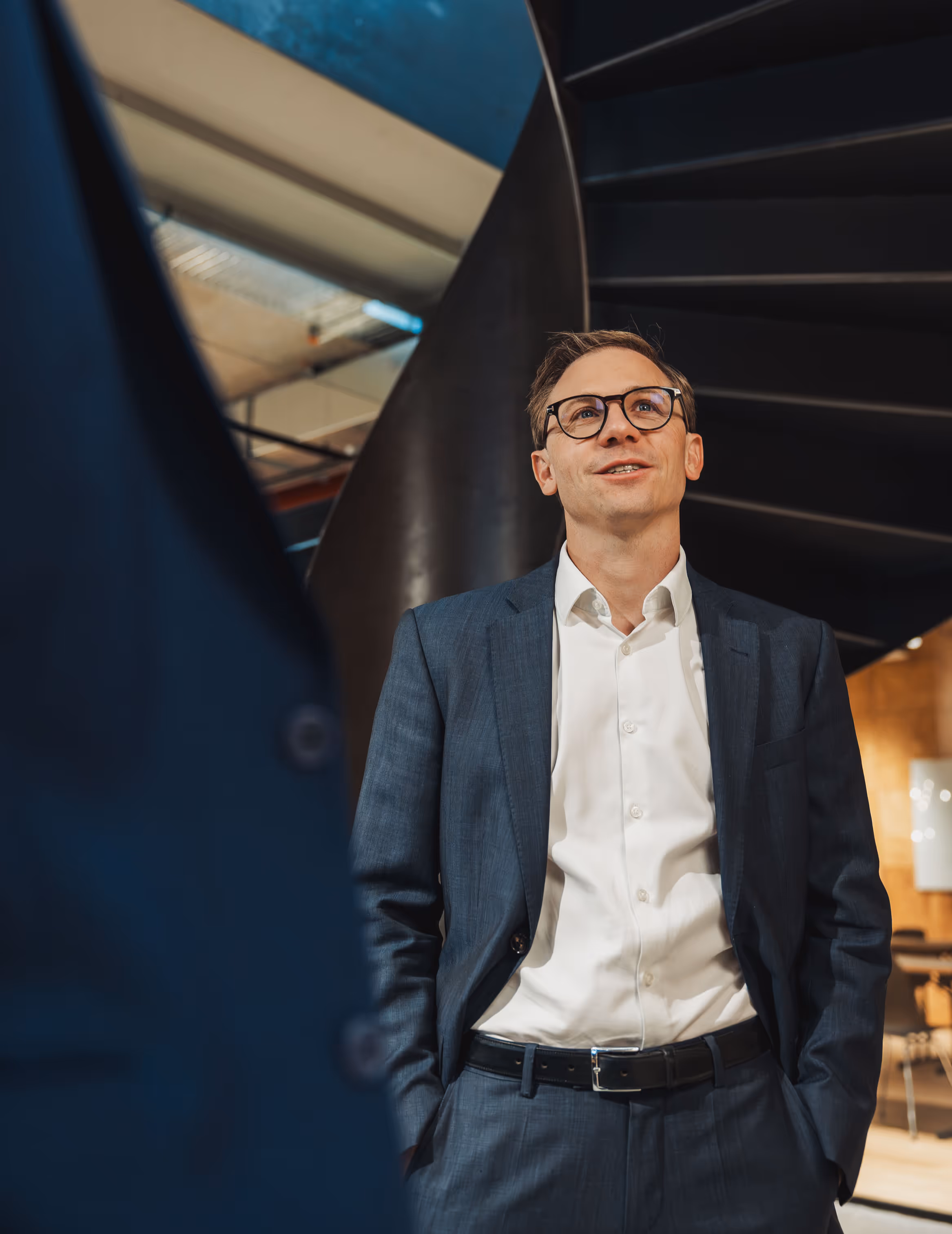 Man in a blue suit and white shirt standing indoors with hands in pockets in front of a black spiral staircase.
