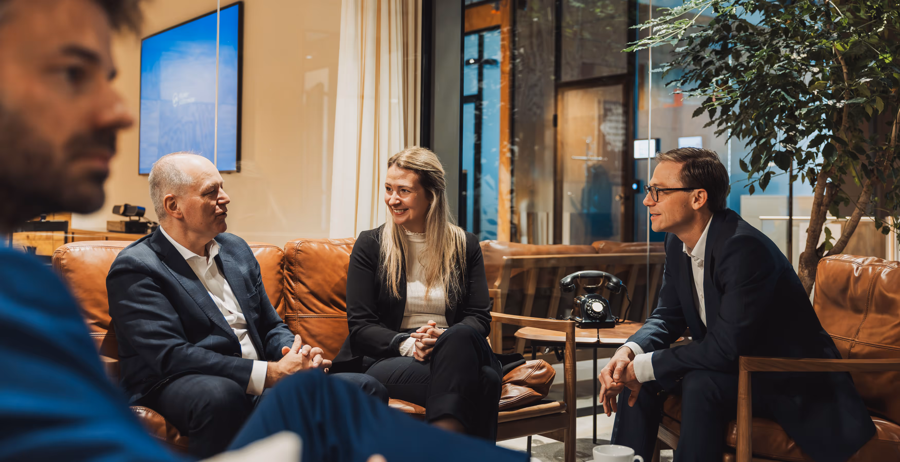 Three business professionals in suits having a discussion while seated on brown leather chairs in a modern office.