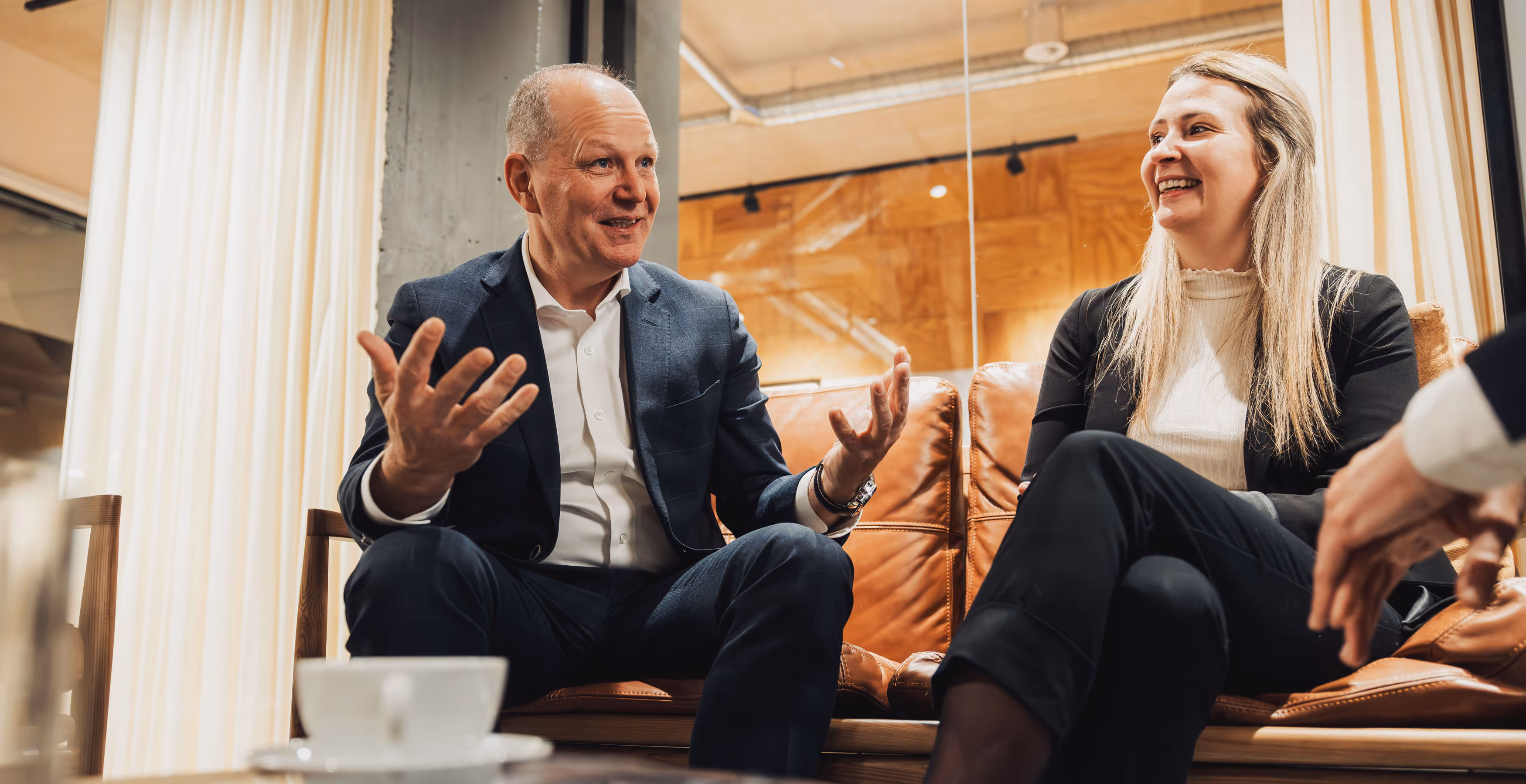 Man in a suit animatedly talking with a woman in business attire sitting on a leather couch in a modern office setting.