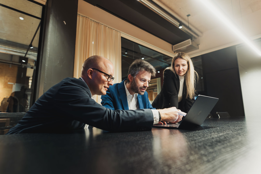 Three business professionals in a meeting, one man in a white shirt speaking with hands on the table while a woman and another man listen attentively.