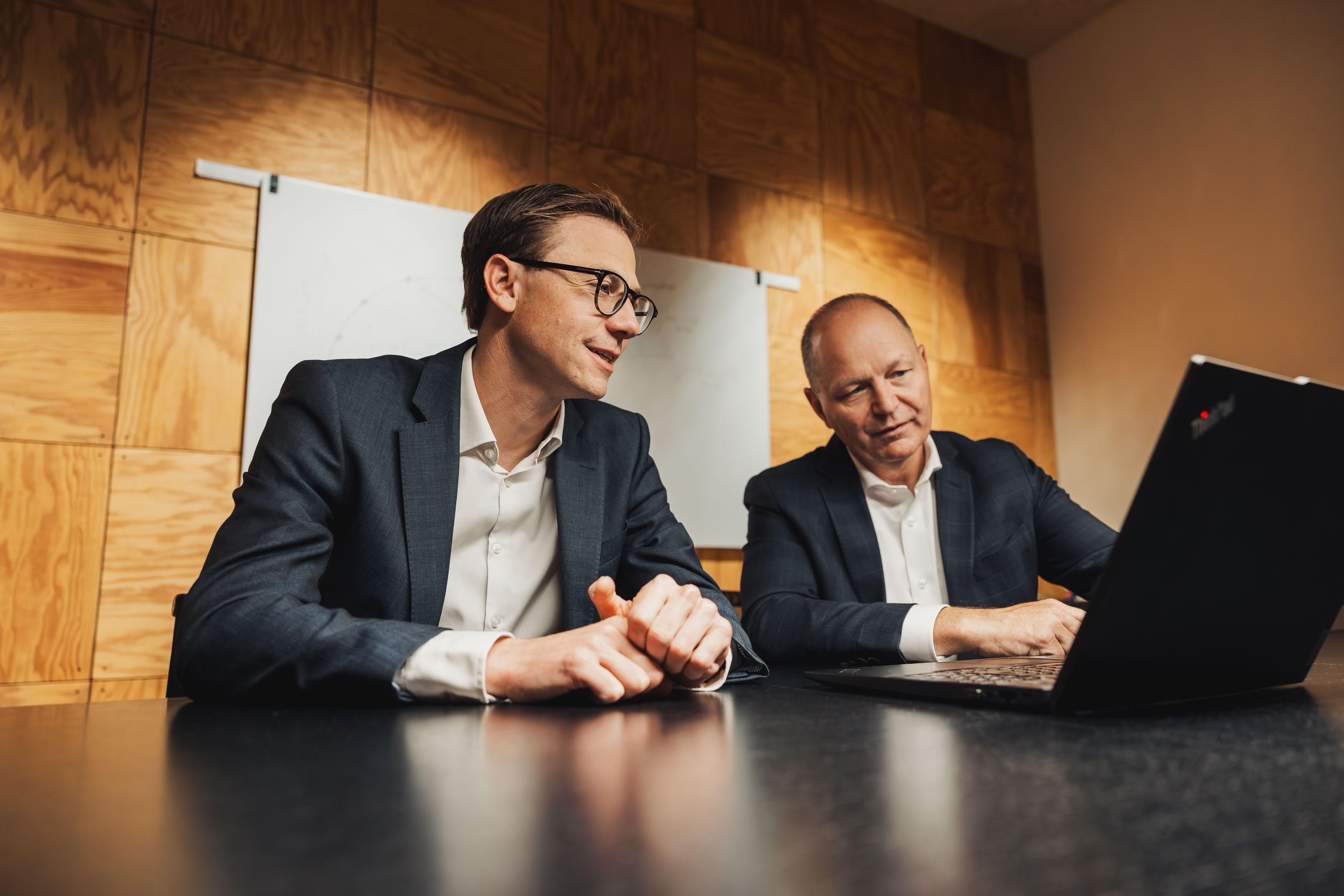 Two businessmen in suits looking at a laptop on a conference table in an office.