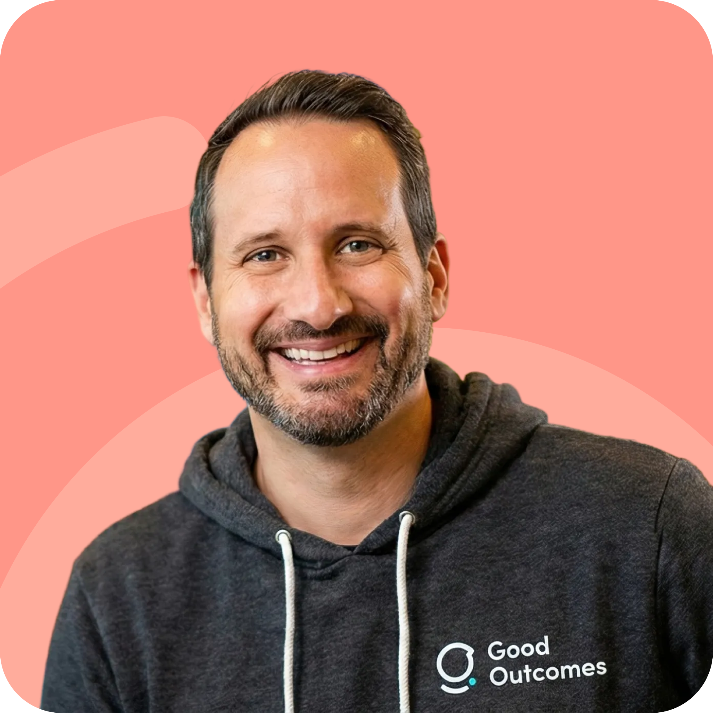Smiling man with short hair and beard wearing a dark hoodie with Good Outcomes logo on a coral background.