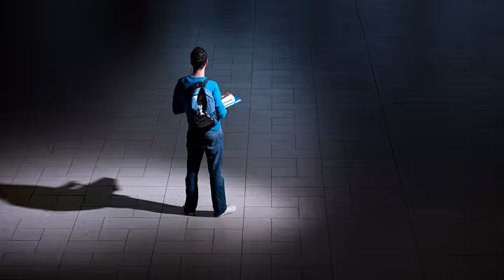 Student standing alone on tiled floor under spotlight holding books with a backpack and casting a long shadow.
