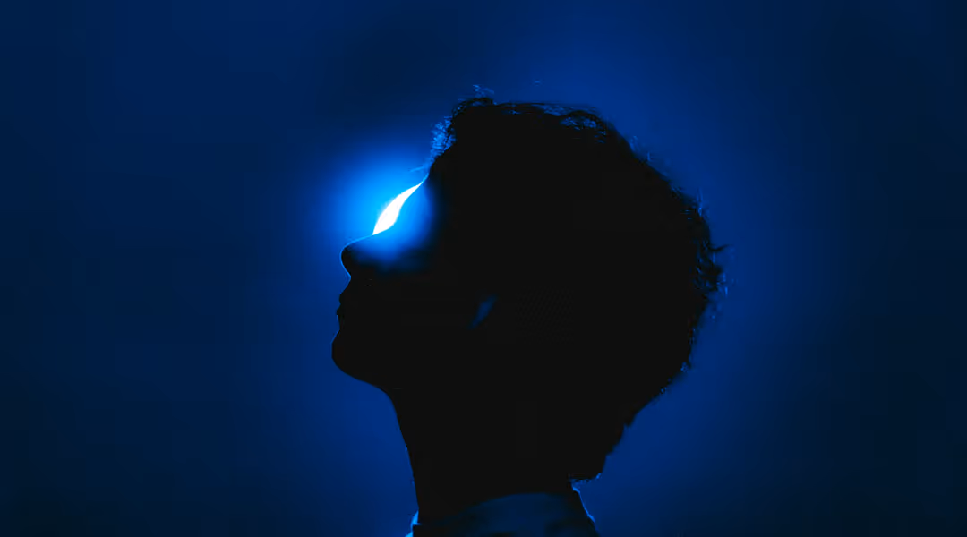 Silhouette of a person in profile with a bright blue light behind their face against a dark blue background.