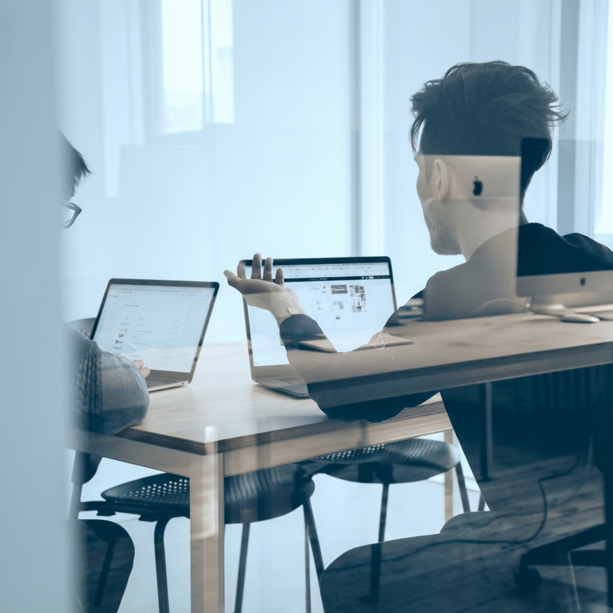 Two people working on laptops and discussing at a table in a modern office with glass walls.