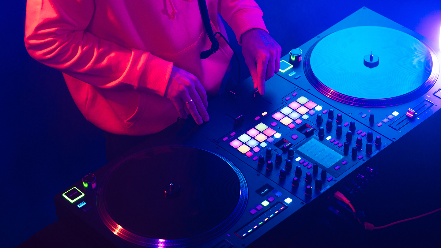 Close-up of a DJ wearing a pink hoodie using a professional DJ controller with lit colorful pads and two turntables, one with a blue vinyl record, in a dimly lit environment with purple and blue lighting.