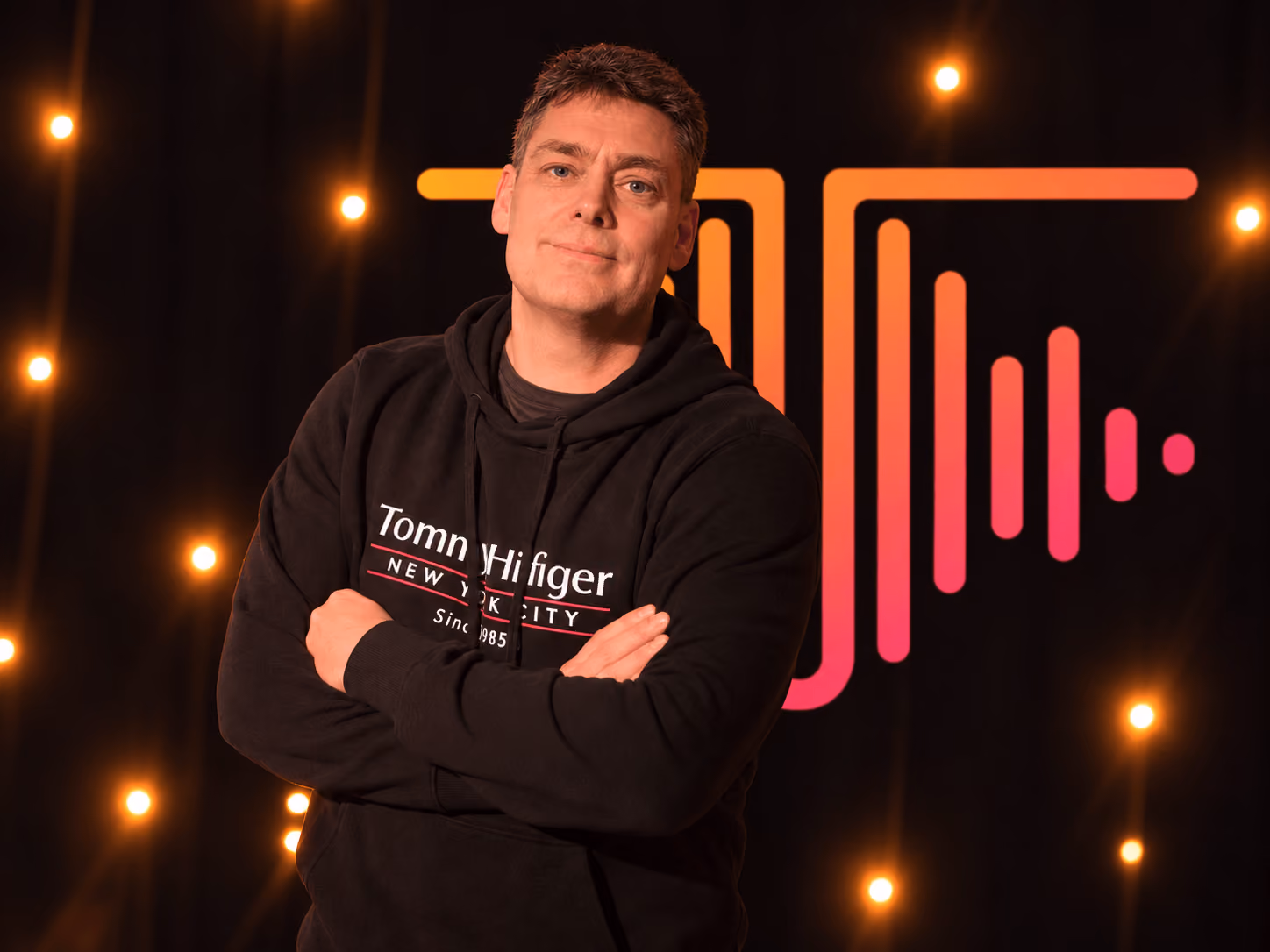 Man with folded arms wearing a black Tommy Hilfiger hoodie, standing against a dark backdrop with orange and pink neon design and warm glowing lights.