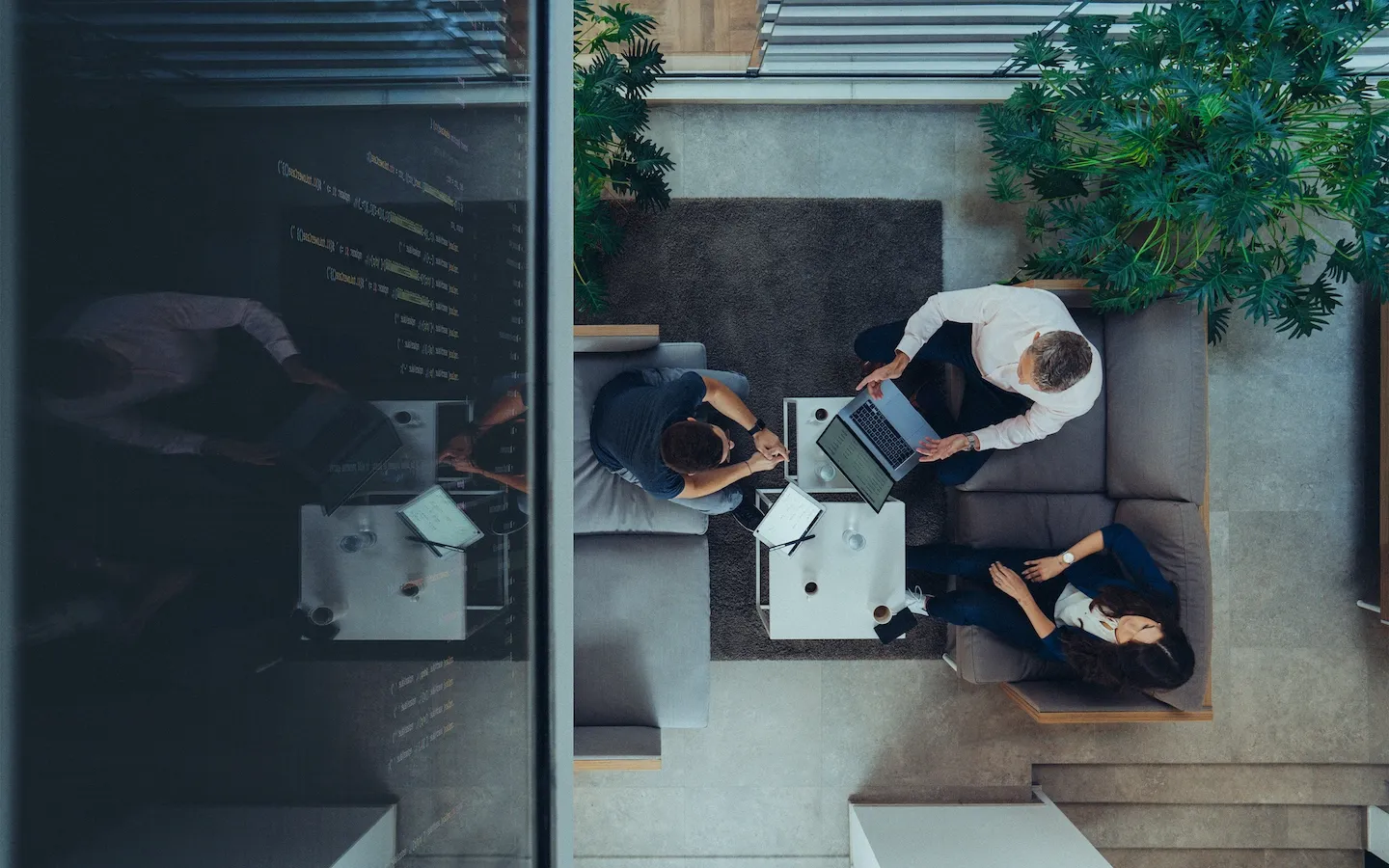 Drei Personen sitzen in einem modernen Büro auf Sofas um einen kleinen Tisch mit Laptop und Notizbüchern, von oben aufgenommen.